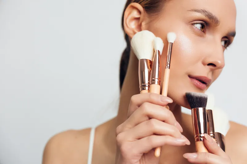 A focused woman applies makeup with brushes near her face, showcasing a detailed beauty routine, professional cosmetics, skincare and grooming tools in a soft studio portrait.