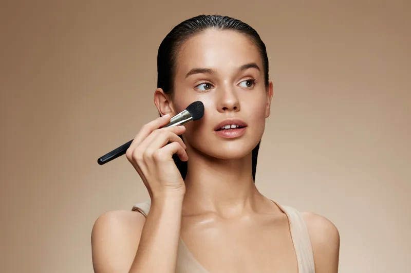 Young woman applying makeup with a brush, showcasing a natural beauty look against a soft beige background, highlighting beauty and self care
