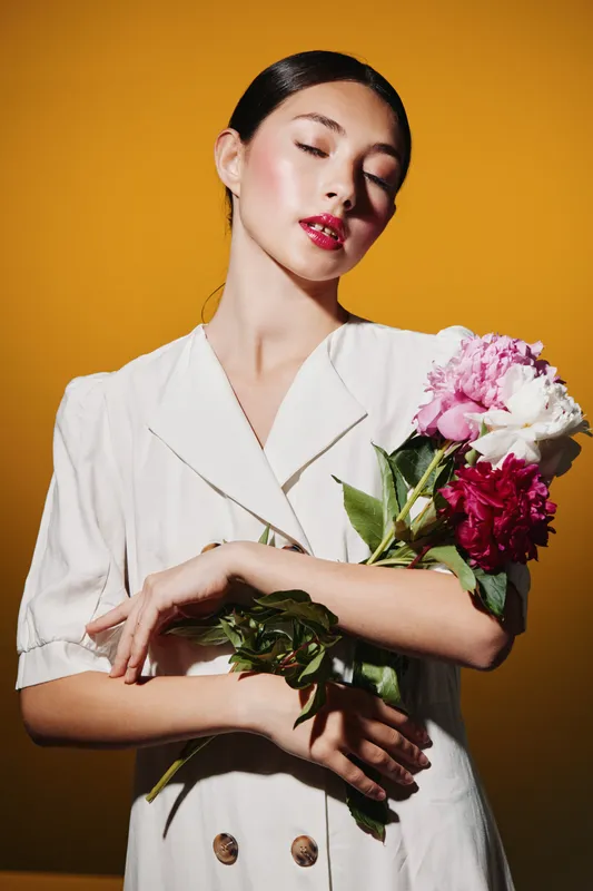 Elegant woman with glossy lips and smooth skin holds vibrant peonies close. She is dressed in crisp white fashion. Studio portrait on a warm yellow background. Beauty concept in fashion and