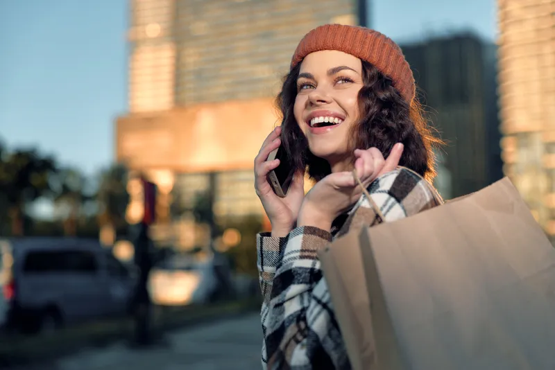 Woman shopping in city with phone and smile, holding a bag in candid moment. Urban portrait evoking authenticity, candid lifestyle, golden hour glow, mindful living and emotional storytelling.