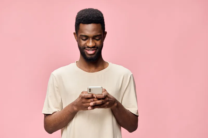 Young African man in a beige shirt smiling while using a smartphone against a pink background, showcasing joy and modern communication