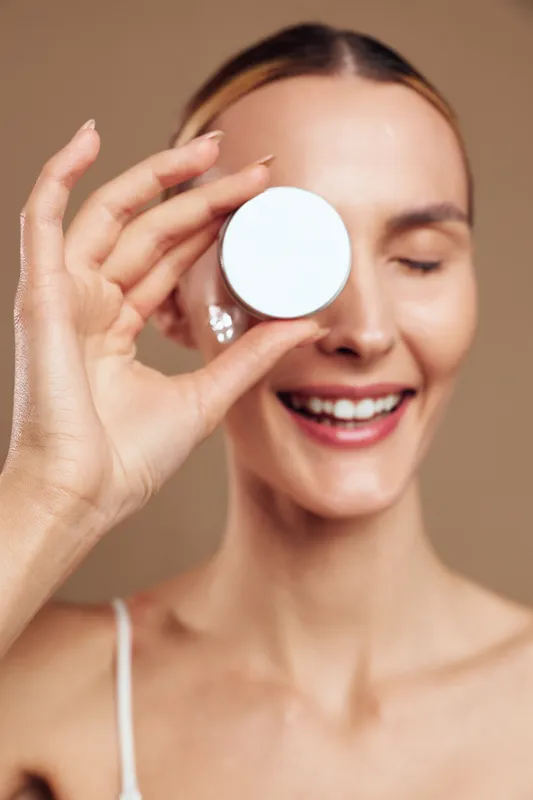 Young woman with clear skin smiles holding a round cosmetic container near her eye isolated on beige background skincare concept beauty and health care product