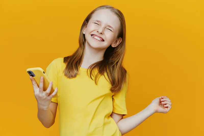 Happy girl smiling with a smartphone against a bright yellow background, wearing a cheerful yellow t shirt, reflecting joy and positivity