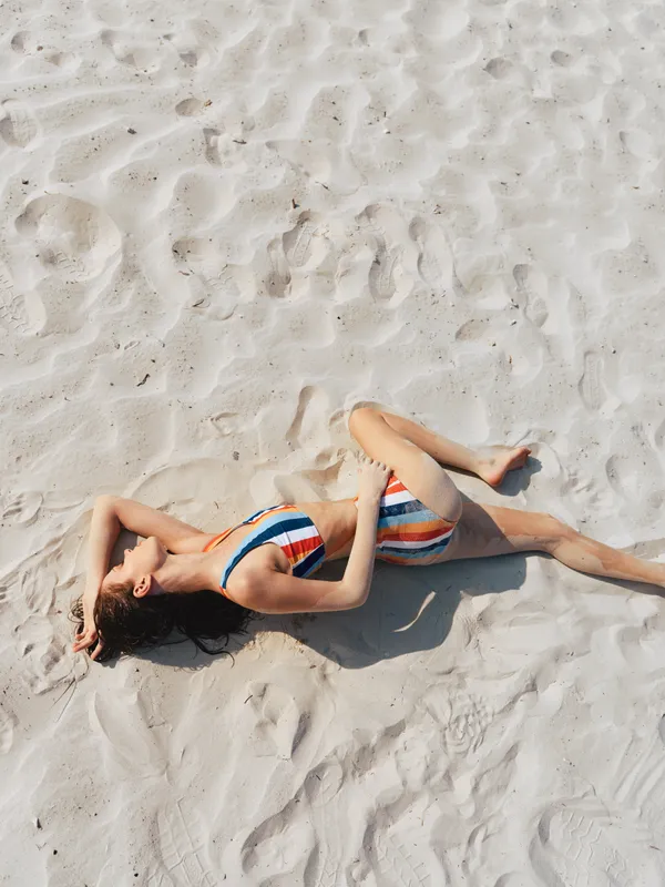A woman lies on soft beach sand in a striped swimsuit, sunlit and relaxed, capturing a serene moment of summer leisure and warm coastal atmosphere.