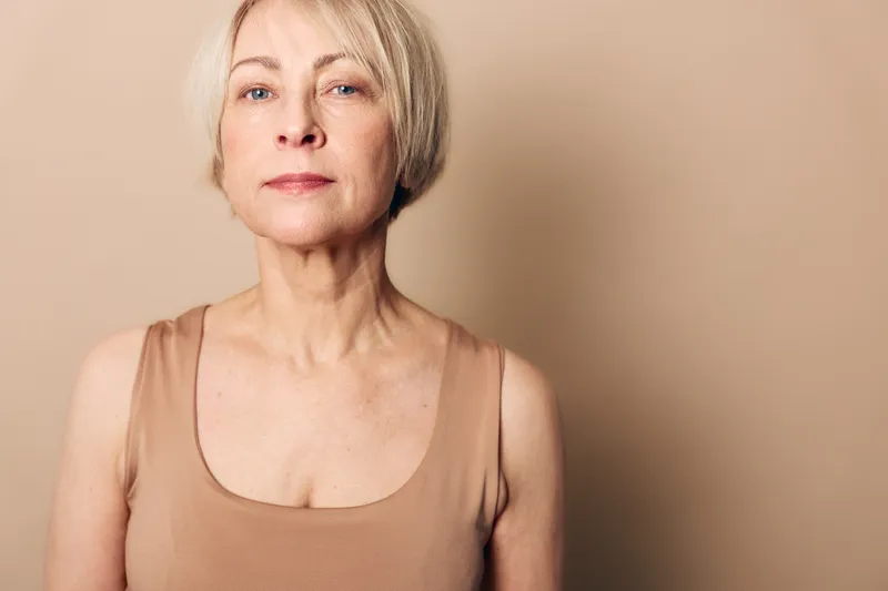 Confident mature woman with short blonde hair wears beige tank top against neutral background. Concept for aging beauty, natural skin, self confidence, and minimalistic portrait photography.