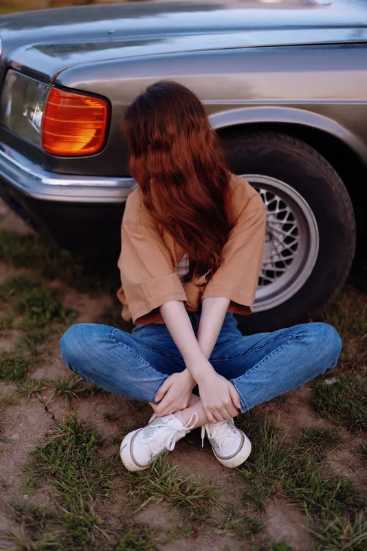 Young woman with long red hair sitting on grass, wearing brown t-shirt and jeans, near vintage car under warm sunlight, evoking casual and relaxed summer vibes.