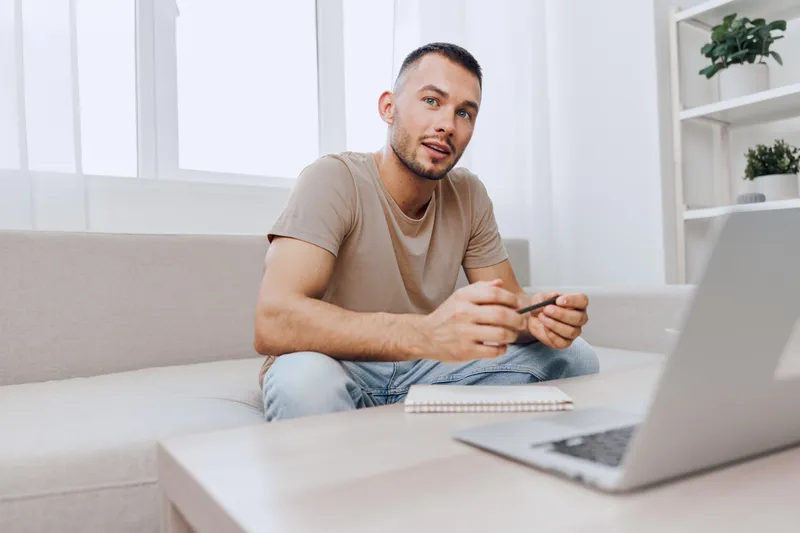 Thoughtful man working at home, engaged in online tasks with a notebook at hand. Modern workspace with minimalistic design and natural light enhances productivity.