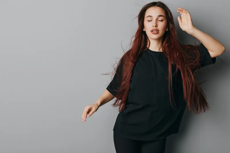 Young woman with long hair wearing black t shirt posing with closed eyes and relaxed expression against gray background in studio. Casual style and peaceful mood.