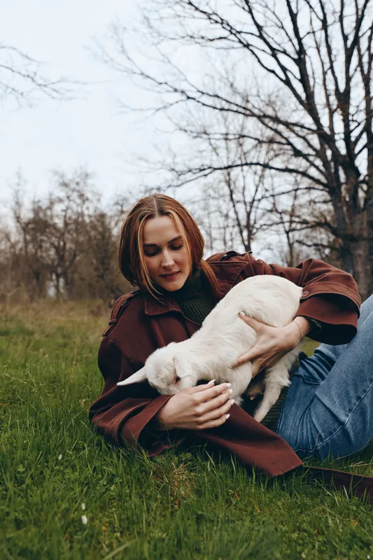Young woman with red hair wearing a brown jacket gently holds a white baby goat while sitting on grass in an autumn park, enjoying a peaceful moment outdoors.
