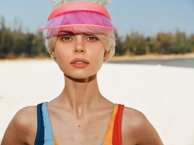 Close portrait of a fashion model on a sunny beach, wearing a multicolor swimsuit and pink visor, highlighting bold summer style and sunlit skin.