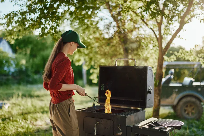 Young woman cooking on a barbecue grill outdoors, dressed in a red shirt and cap, enjoying a sunny summer day in nature, showcasing joyful emotions and vibrant colors