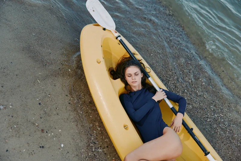 A solitary person relaxes in a yellow kayak along a quiet shoreline, resting under a calm sky as gentle waves touch the sand, bright tones inviting pause.