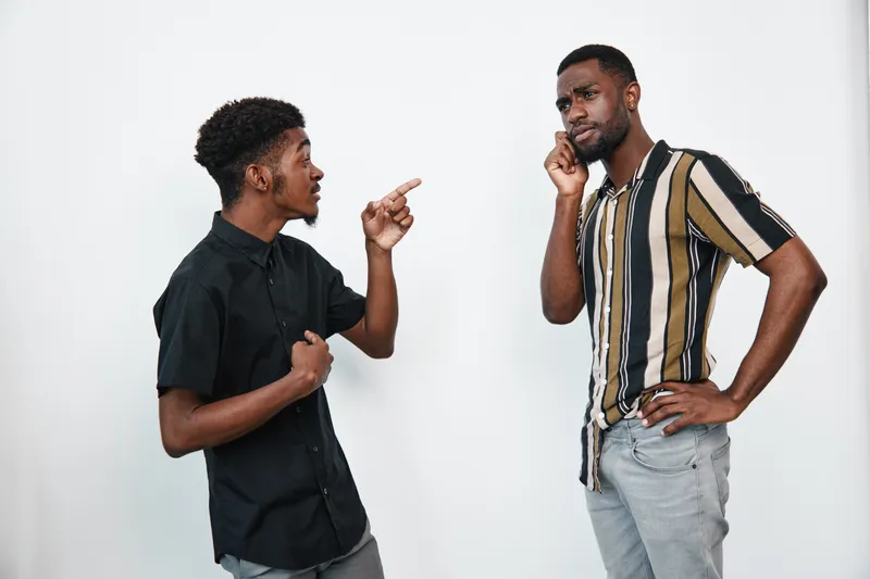 Two black men with dark skin having a serious conversation in casual clothes on white background. One man is pointing while the other listens thoughtfully with hand on chin.