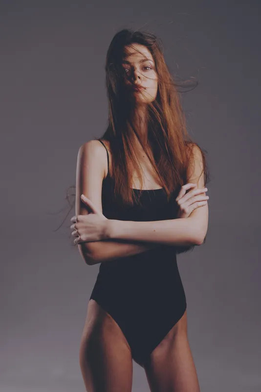 slender woman in black bodysuit with loose hair and neutral expression, minimalistic dark background, fashion portrait concept