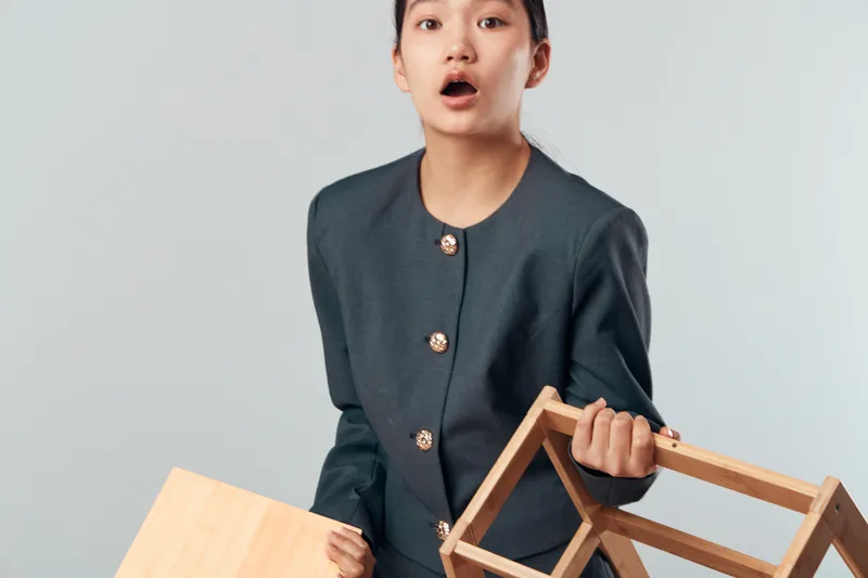 Young woman in dark jacket holding wooden box and frame with surprised expression on neutral background. Studio portrait with casual clothing and unexpected emotion captured.