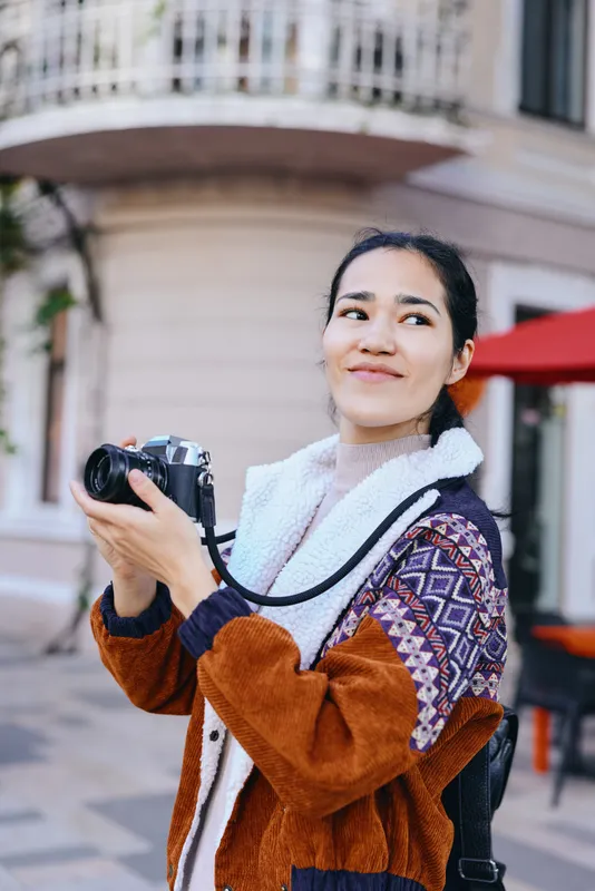 Camera, photography, woman, outdoors, street, urban, lifestyle, smile, jacket, scarf capture a candid moment as she holds a camera, exploring a sunny plaza with colorful surroundings and a cheerful