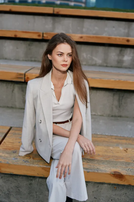 Woman in white business outfit sitting on wooden steps with thoughtful expression, wearing a light blazer and trousers, outdoor urban setting, fashion theme