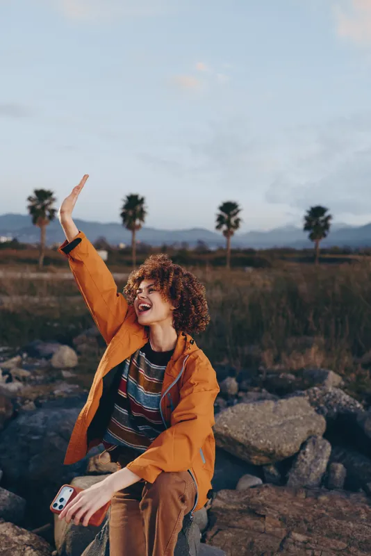 Woman lifestyle outdoors, smiling, curly hair, orange jacket, sunset, palm trees, rocks, raising one arm on a rocky overlook, striped top, warm golden light, relaxed joyful moment