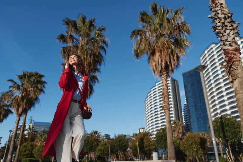 Young woman in a long red coat walks confidently along a sunny urban street lined with palm trees and modern high-rise buildings under a clear blue sky.