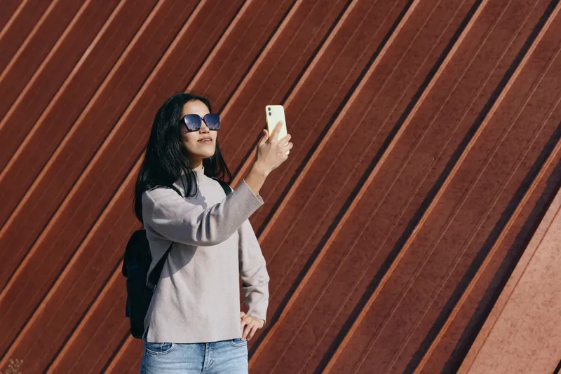 Selfie of a woman wearing sunglasses and a light sweater, holding a smartphone against a rusted diagonal panel wall, casual urban style, daylit fashion candid