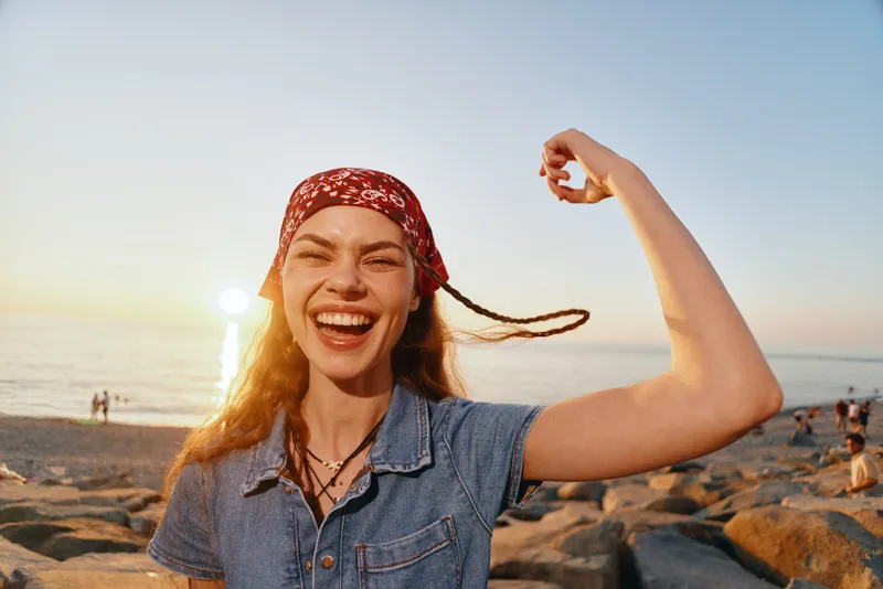 Lifestyle woman in modern boho-western denim street style smiling joyfully by the beach during golden hour with film color warmth and relaxed summer vibe.