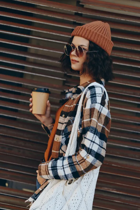 Woman with coffee in urban fashion outfit wearing beanie, sunglasses and tote, candid lifestyle portrait at golden hour glow. Authenticity, mindful living and emotional storytelling mood.