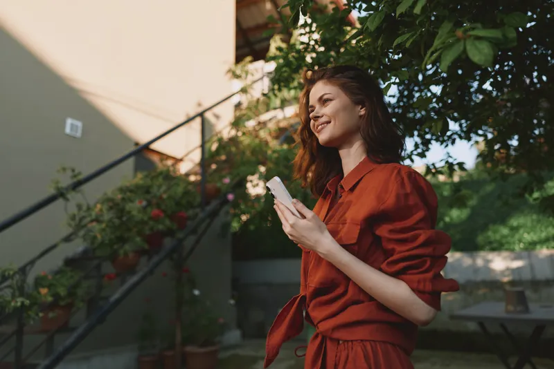 A young woman wearing a rust dress smiles while standing outdoors, gently holding a phone as warm sunlight lights a leafy courtyard, creating a relaxed lifestyle moment