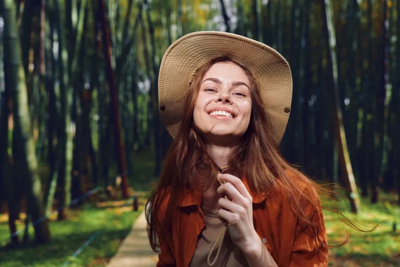 Woman smiling in a straw hat on a forest path, portrait of a happy young woman enjoying nature. Outdoors summer scene with carefree expression, sunlight and blurred trees background.