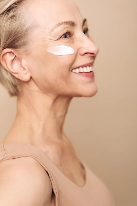 Smiling mature woman with short hair applies white face cream on cheek in beige top. Close-up skincare concept for healthy skin, anti-aging, beauty routine and natural wellness in studio light.
