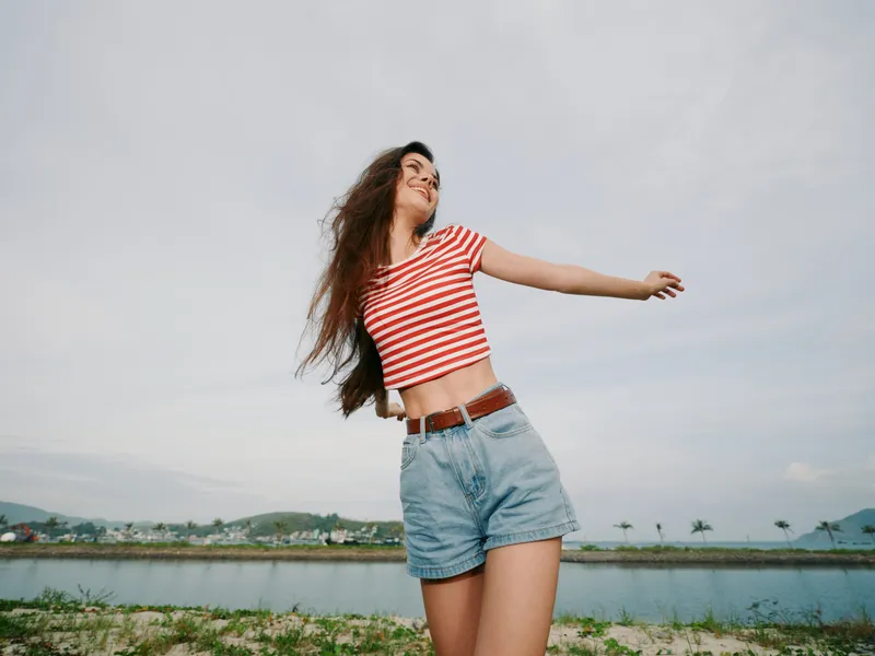 Young woman stands with arms outstretched in a breezy striped crop top and denim shorts, radiating joy under a bright sky with expansive landscape and distant hills.