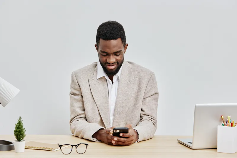 Professional young African American man with short curly hair and beard, wearing a beige blazer over a white shirt, sitting at a minimalist office desk, using smartphone, with laptop, glasses, and pot