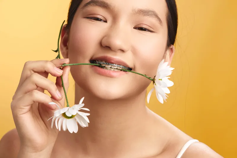 asian woman smiles softly as daisies are held between her lips, creating a playful yet serene portrait against a warm, bright backdrop; her cheerful expression exudes freshness, optimism, and natural