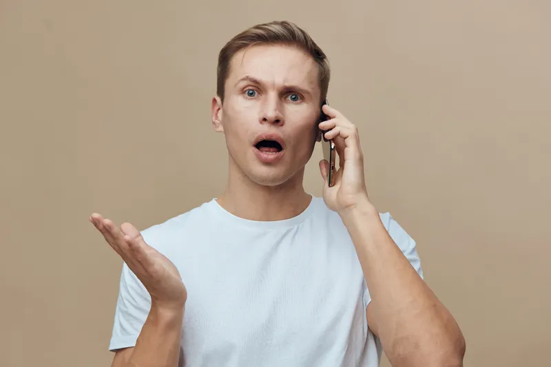 Confused young Caucasian man in casual t-shirt talking on mobile phone with puzzled expression, hand raised in question, isolated on beige background, studio portrait, human emotions, communication co