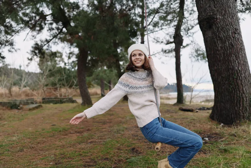 Swinging woman in a cozy knit sweater and jeans enjoys an outdoor moment in a park, with tall trees and soft natural light creating autumn mood.