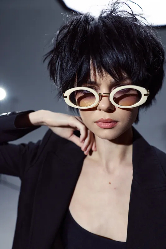 Stylish woman with short black hair wearing round white sunglasses and dark jacket posing confidently in studio light.