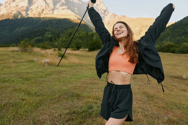 A joyful young woman poses outdoors in a sunny landscape, stretching her arms with a wide smile, wearing casual athleticwear, exuding energy and freedom in nature.