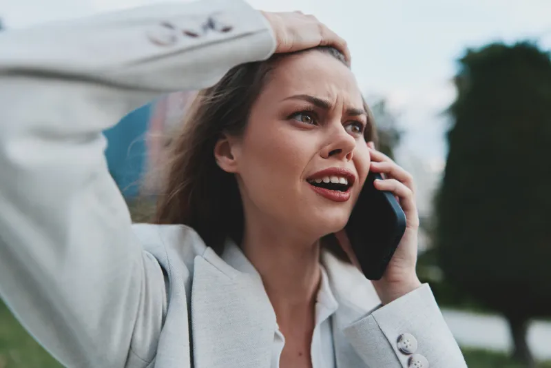Stressed woman talking on phone outdoors showing financial crisis emotions in casual business outfit with blurred natural background