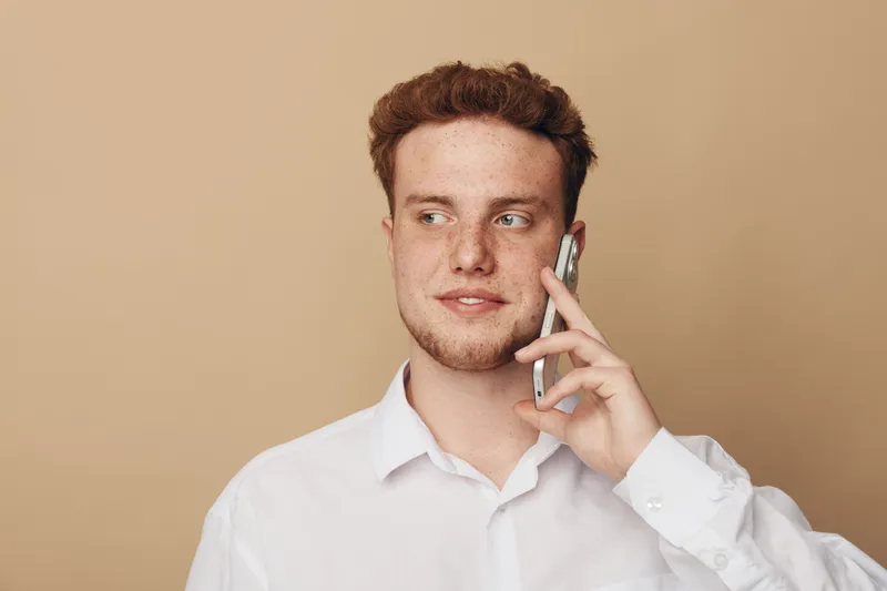 Young man with red hair wearing white shirt talks on smartphone looking sideways. Isolated on beige background. Concept of communication, technology, and casual lifestyle.