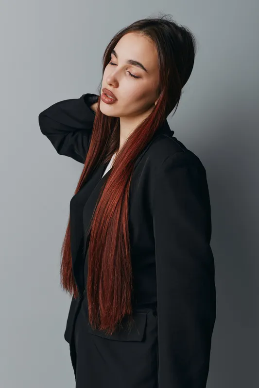 Young woman with long braided hair posing with closed eyes and relaxed expression wearing black jacket and white shirt against plain gray background in studio.