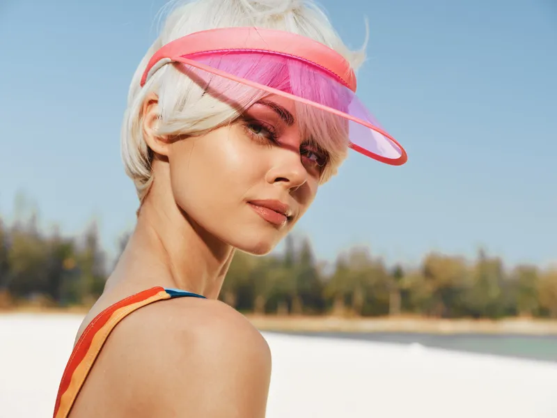 Portrait of a blonde woman wearing a pink visor on a sunny beach, with glowing skin and a calm expression, set against a bright sky and soft sandy backdrop.