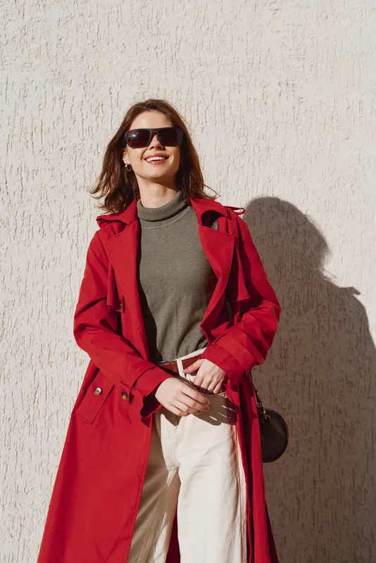 Cheerful woman outdoors in a bold red coat, sunglasses, and olive sweater, standing by a sunlit wall, relaxed pose, warm tones, natural light, carefree mood