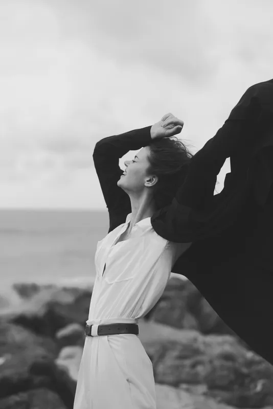 Woman in a white dress on a windy coast, lifting a dark cape to express freedom, movement and dramatic fashion in monochrome, cinematic lighting and calm sea backdrop