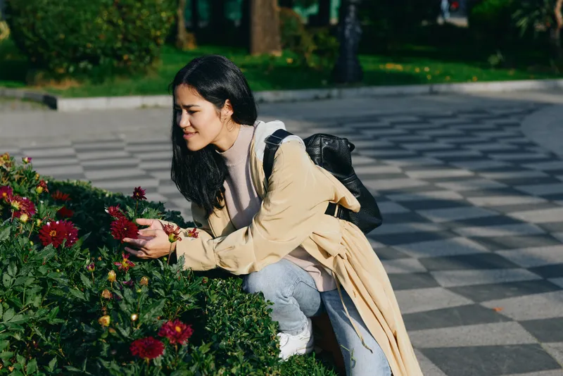 Woman kneels beside vibrant flowers in a park, observing blossoms up close, wearing a beige coat and backpack, enjoying a calm outdoor moment among greenery.