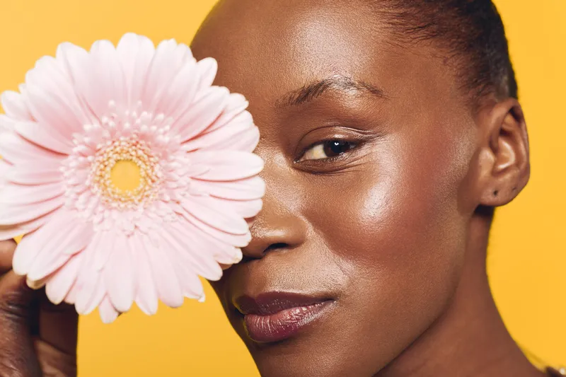 Close-up of African woman with dark skin holding pink gerbera flower near her eye. Bright yellow background highlights natural beauty and floral concept for skincare and wellness.