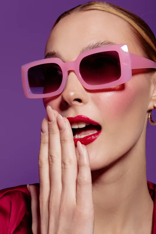 Close-up of a fashionable woman wearing pink sunglasses and red lipstick, covering her mouth with her hand. Bright makeup and purple background create a stylish, modern look.