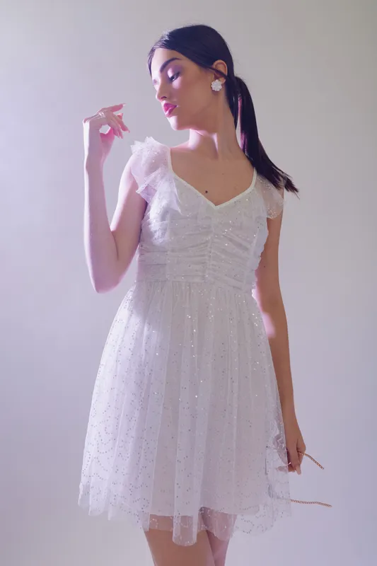 Young woman in a white dress poses elegantly with a side glance. Soft lighting highlights her beauty and fashion style. Studio shot with a light background.