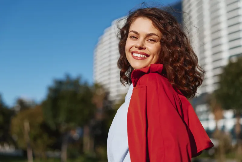 A confident young woman with curly hair wears a red cape and smiles at the camera in an urban park, with modern buildings in the background on a sunny day, radiating energy.