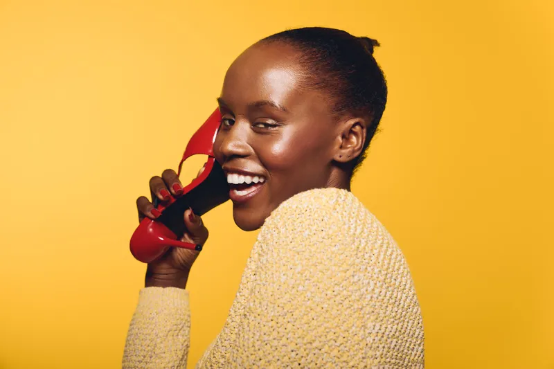 Happy African woman with dark skin wearing yellow sweater holds red high heel shoe near face smiling on yellow background. Concept for fashion, beauty, joy, and lifestyle portrait.