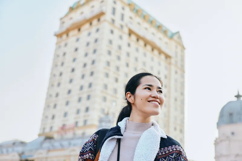 Woman with a warm smile stands outdoors beside a tall city building, wearing a knit sweater and scarf, captured against an urban architectural backdrop during daytime Bright sky and breeze today.