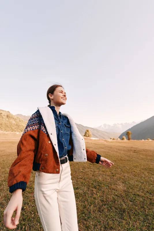 Woman walking in a grassy field near distant mountains, wearing a cozy jacket and denim shirt, smiling as she explores autumn landscape and fresh air in a peaceful outdoor lifestyle scene.